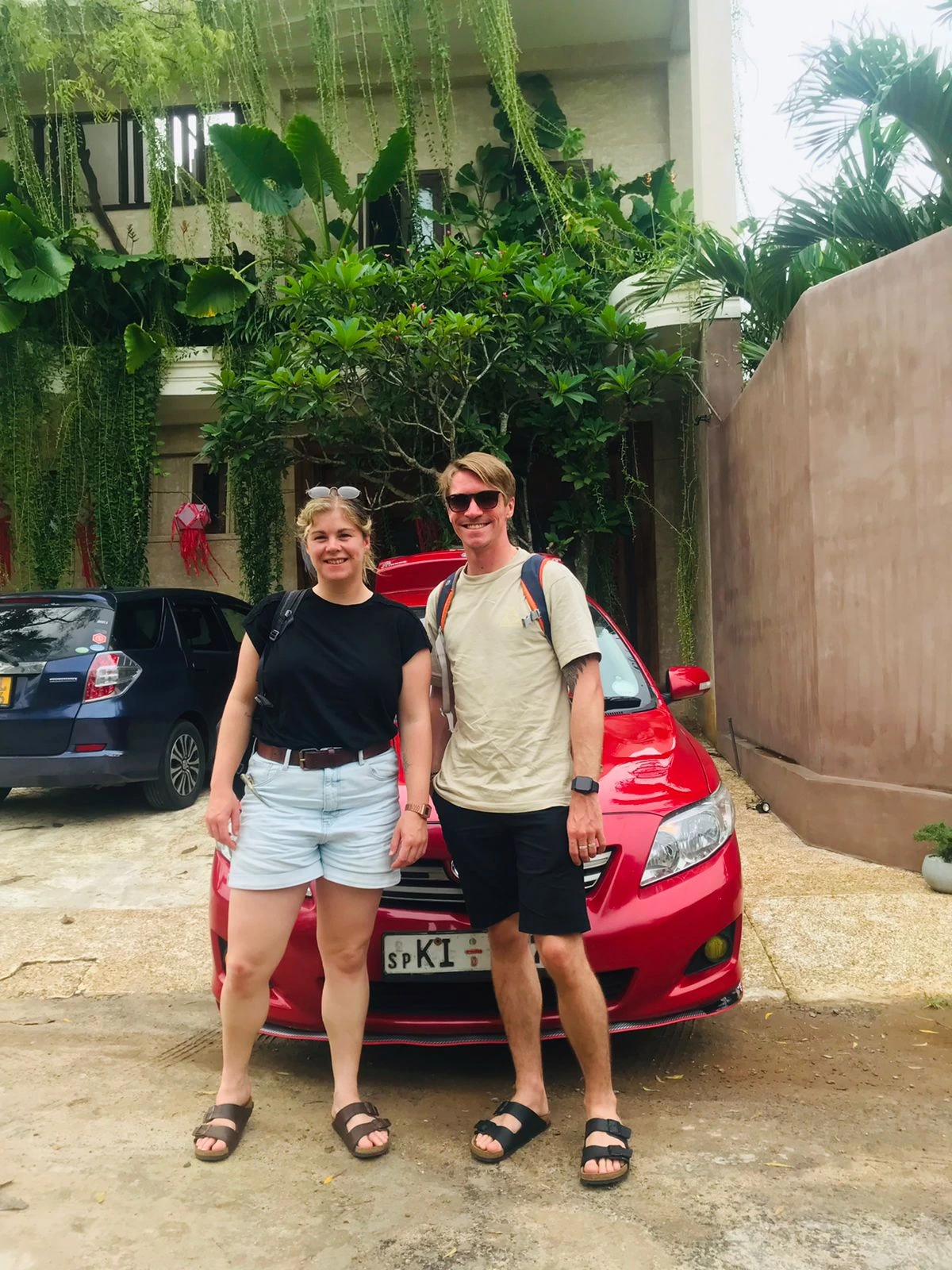 Couple tourists with red sedan at guesthouse gate Sri Lanka - Young travelers standing by comfortable tour car during accommodation stop on island tour