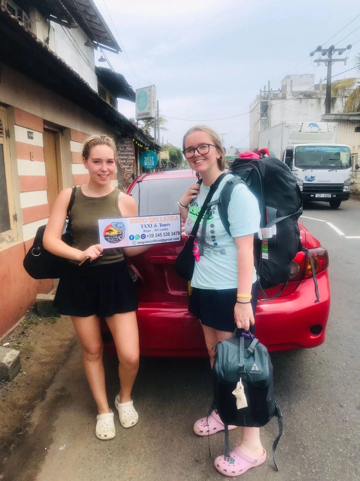 Two female tourists with red tour car Sri Lanka - Young women travelers holding Wego Taxi Tours sign standing next to comfortable sedan during road trip